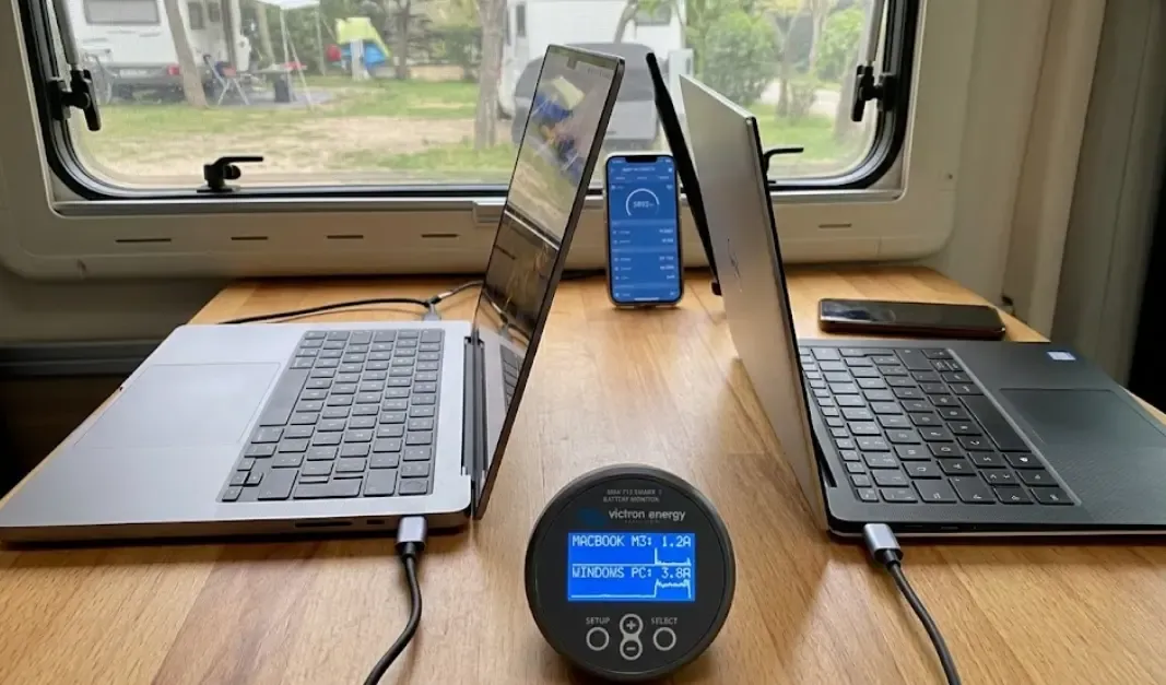 Comparativa de un MacBook M3 y un portátil Windows en una mesa camper, con un monitor de batería Victron mostrando la diferencia de amperios consumidos en tiempo real.