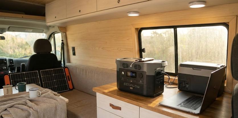 Interior de furgoneta camper equipada con una estación de energía portátil sobre la encimera alimentando un ordenador portátil y una nevera de compresor, con paneles solares plegables al fondo, demostrando una solución sin instalación eléctrica fija.